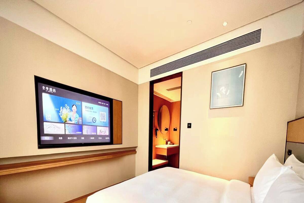 A hotel room with a bed, a TV mounted on the wall, a small table, and a bathroom visible through an open door.
