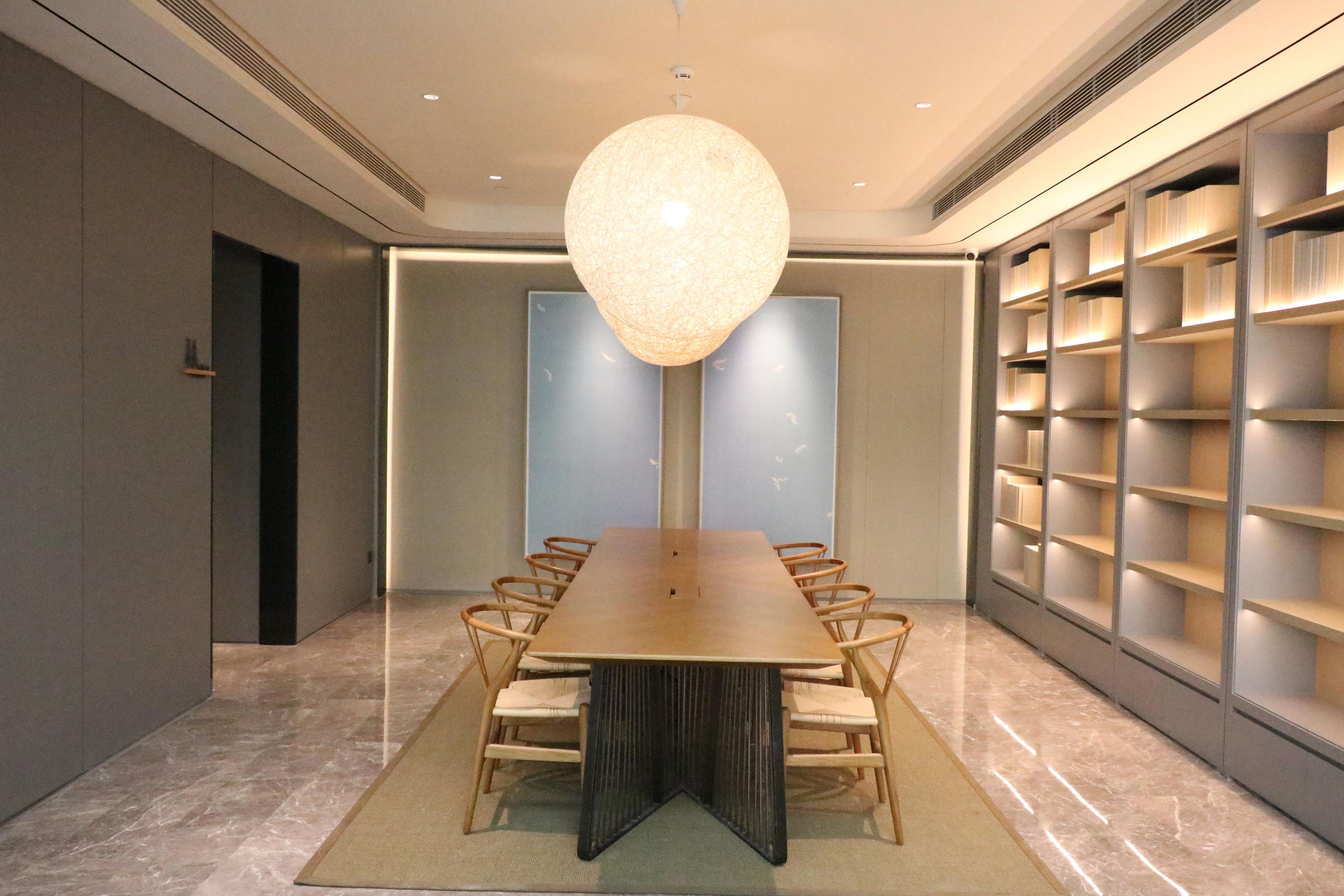 A conference room with a long table, chairs, a central hanging light fixture, and bookshelves along the walls.