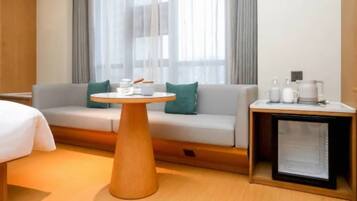 A hotel room with a sofa, a round wooden table, a bedside table with a lamp, and a window with curtains.