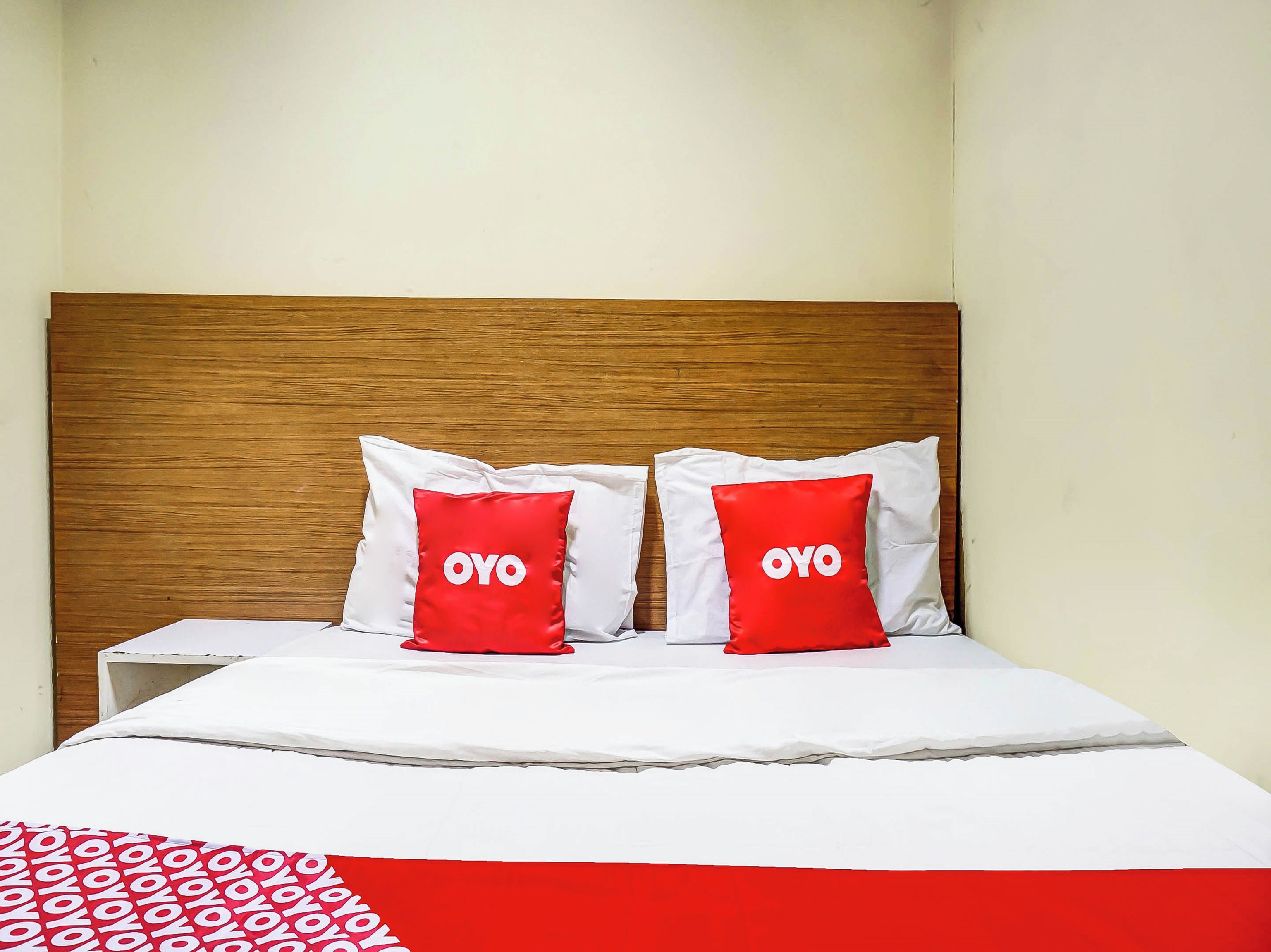 Un lit avec une tête de lit en bois, deux oreillers rouges ornés du mot « OYO », une literie blanche et une couverture à motifs rouges et blancs.