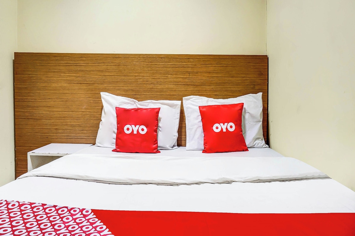 Una cama con cabecera de madera, dos almohadas rojas con el texto 'OYO', ropa de cama blanca y una manta estampada en rojo y blanco.