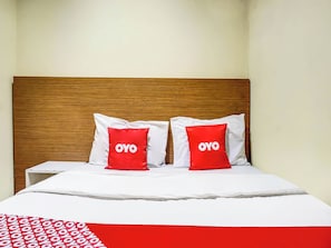 Una cama con cabecera de madera, dos almohadas rojas con el texto 'OYO', ropa de cama blanca y una manta estampada en rojo y blanco.