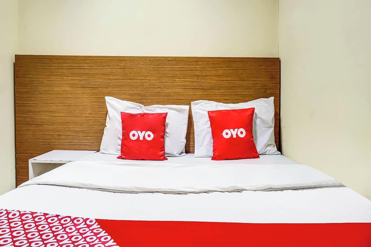 Een bed met een houten hoofdbord, twee rode kussens met de tekst 'OYO', witte beddengoed en een rood-wit gebloemd plaid.