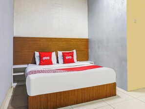 Una habitación de hotel con cabecera de madera, cama con ropa de cama blanca y almohadas rojas, una manta roja y una mesita de noche blanca.