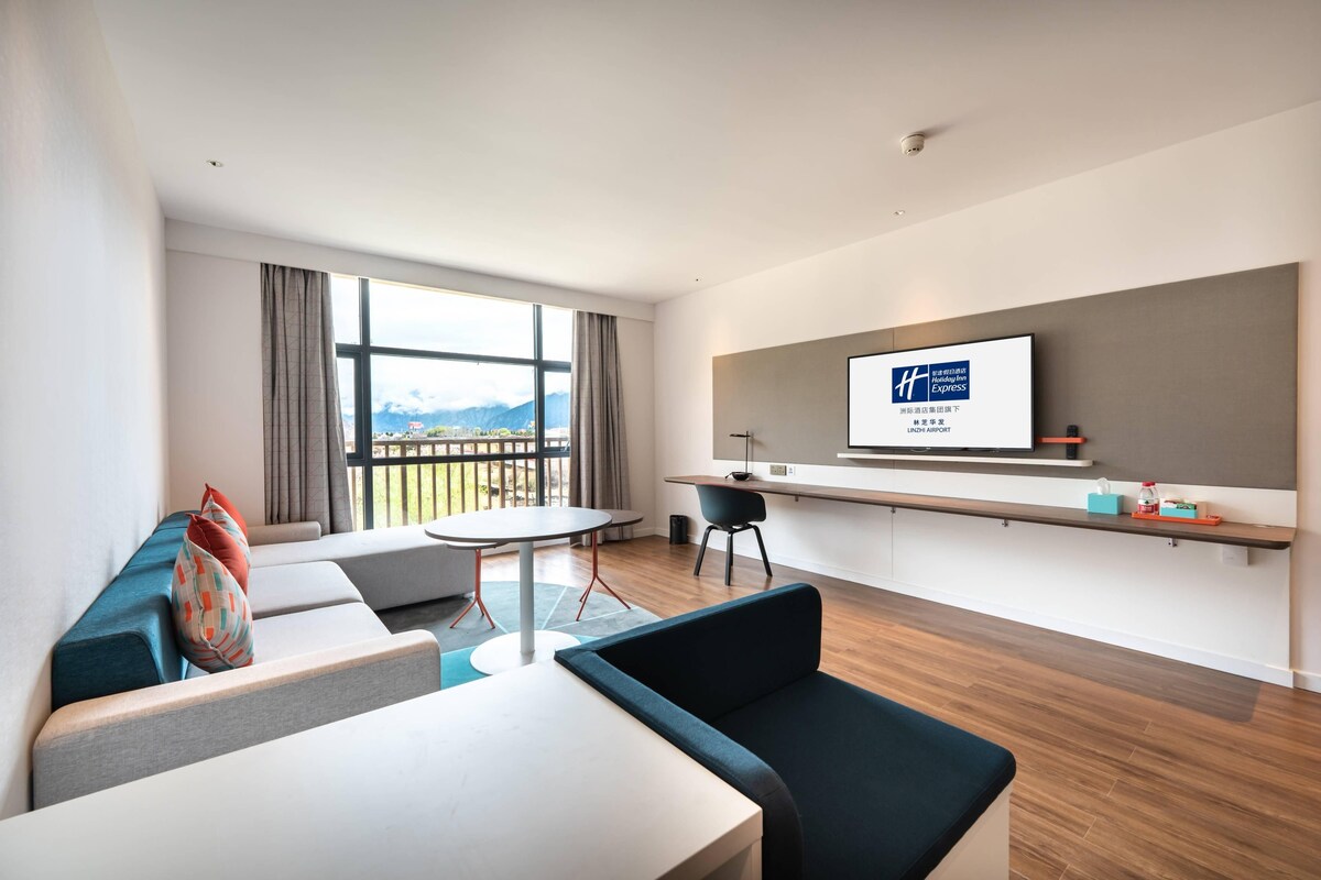 A modern hotel room with a large window, a flat-screen TV, a sofa, a dining table, and a small kitchen area.