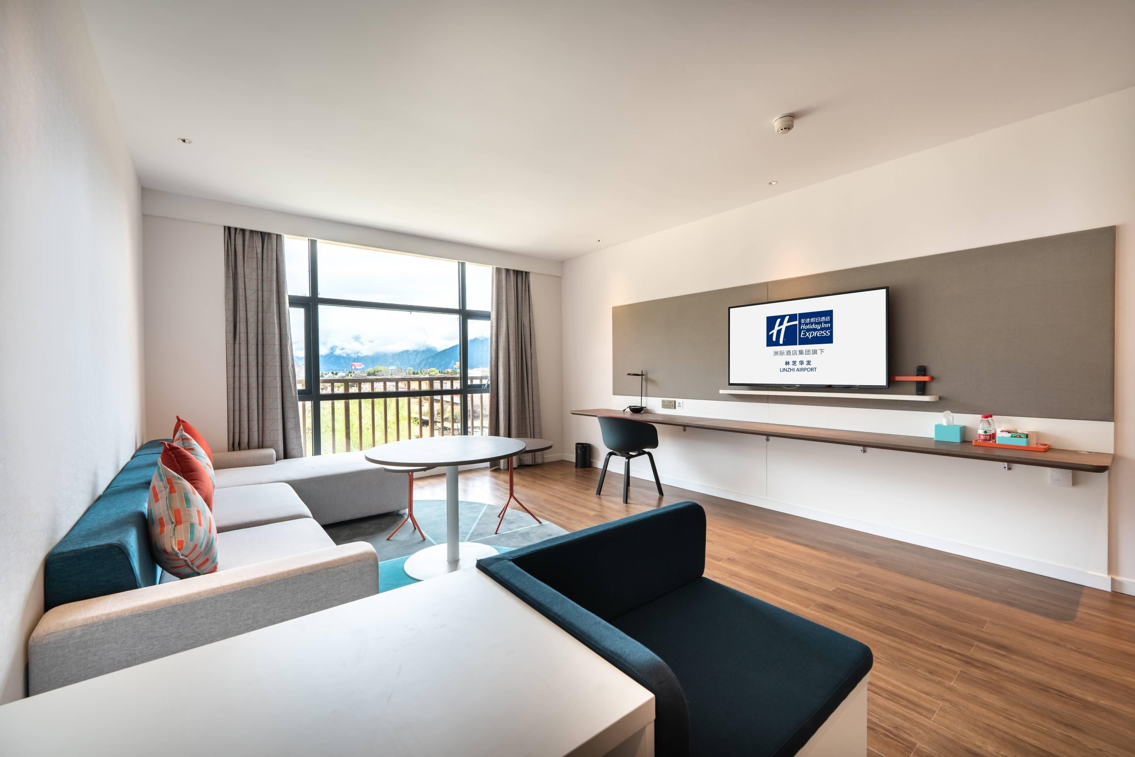A modern hotel room with a large window, a flat-screen TV, a sofa, a dining table, and a small kitchen area.