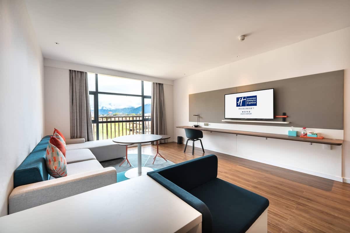 A modern hotel room with a large window, a flat-screen TV, a sofa, a dining table, and a small kitchen area.
