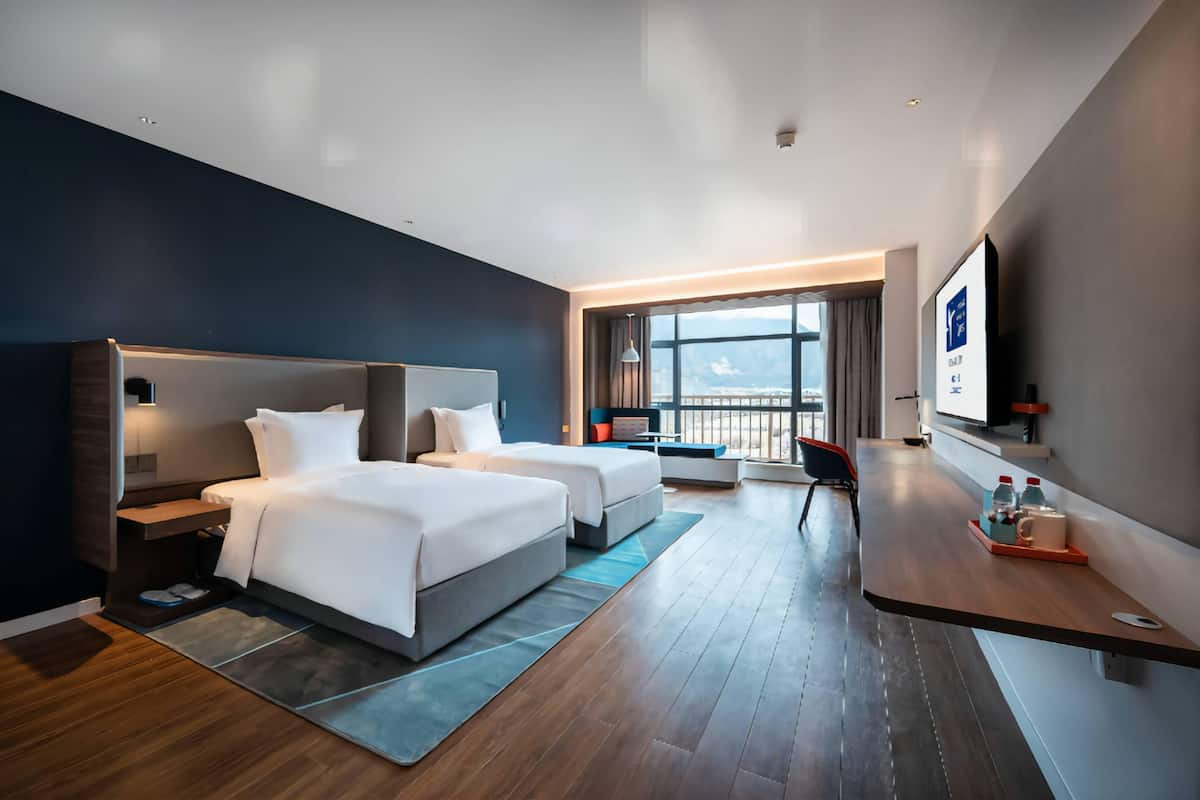 A modern hotel room with two beds, a desk, a TV, and a balcony with a view.