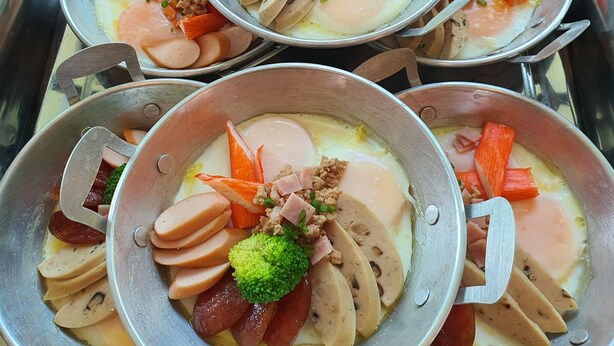 Disposición de buffet de acero inoxidable con múltiples recipientes que contienen carnes, verduras y huevos en rodajas.