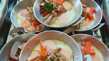 Disposición de buffet de acero inoxidable con múltiples recipientes que contienen carnes, verduras y huevos en rodajas.