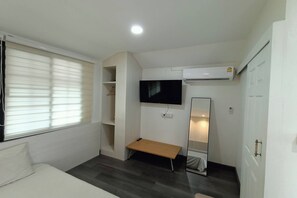 Une petite chambre à coucher moderne, équipée d’un lit, d’un banc, d’une télévision, d’un climatiseur et d’un miroir.
