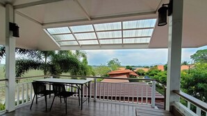 Un espacio al aire libre techado con una mesa y sillas, vista a zonas verdes y edificios a lo lejos, y una barandilla de metal.