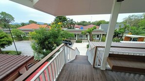Una escalera de madera conduce a un espacio al aire libre techado, con vistas a edificios residenciales y vegetación.