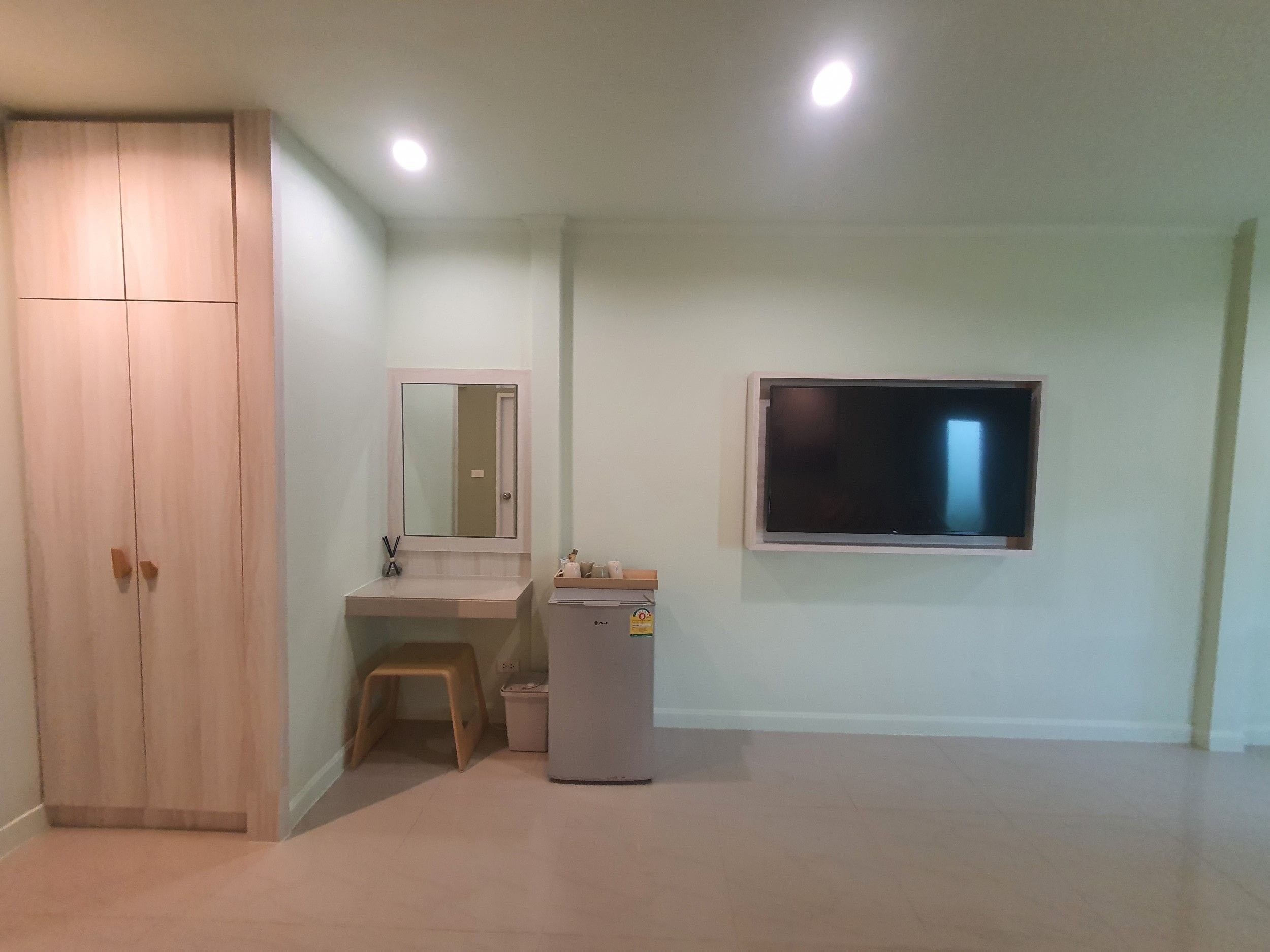 Une pièce comprenant un placard, un bureau avec un miroir, un petit tabouret, une poubelle et un téléviseur fixé au mur.