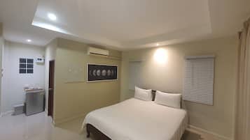 A bedroom with a bed, a wall-mounted air conditioner, a window with blinds, and a door leading to another room.