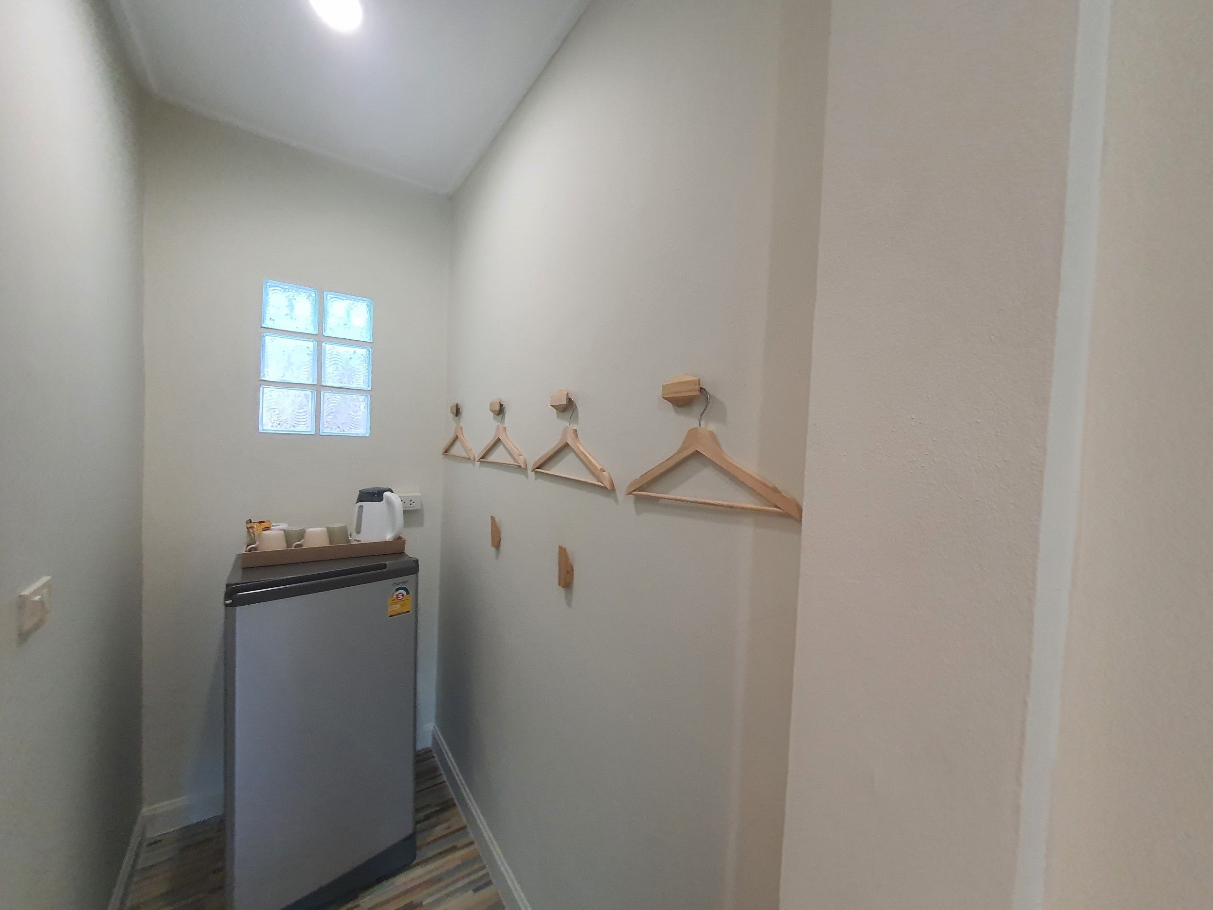 A narrow hallway with a refrigerator, wooden hangers on the wall, and a window with blinds.