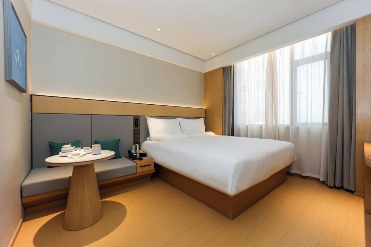 Quarto de hotel moderno com uma cama grande, uma mesa redonda de madeira, área de estar com almofadas e uma janela ampla com cortinas.