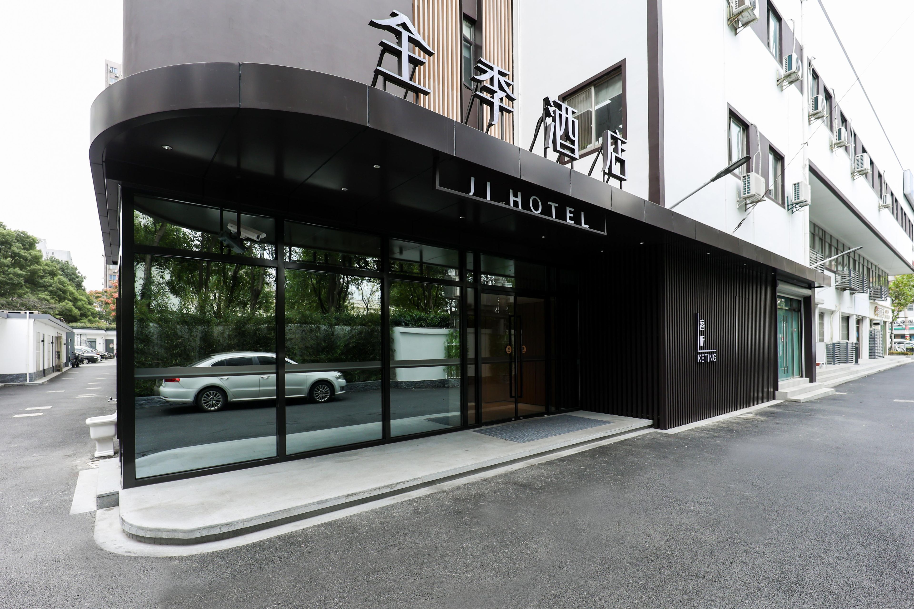 A modern hotel entrance with a glass facade, a car parked outside, and a clear view of the street.