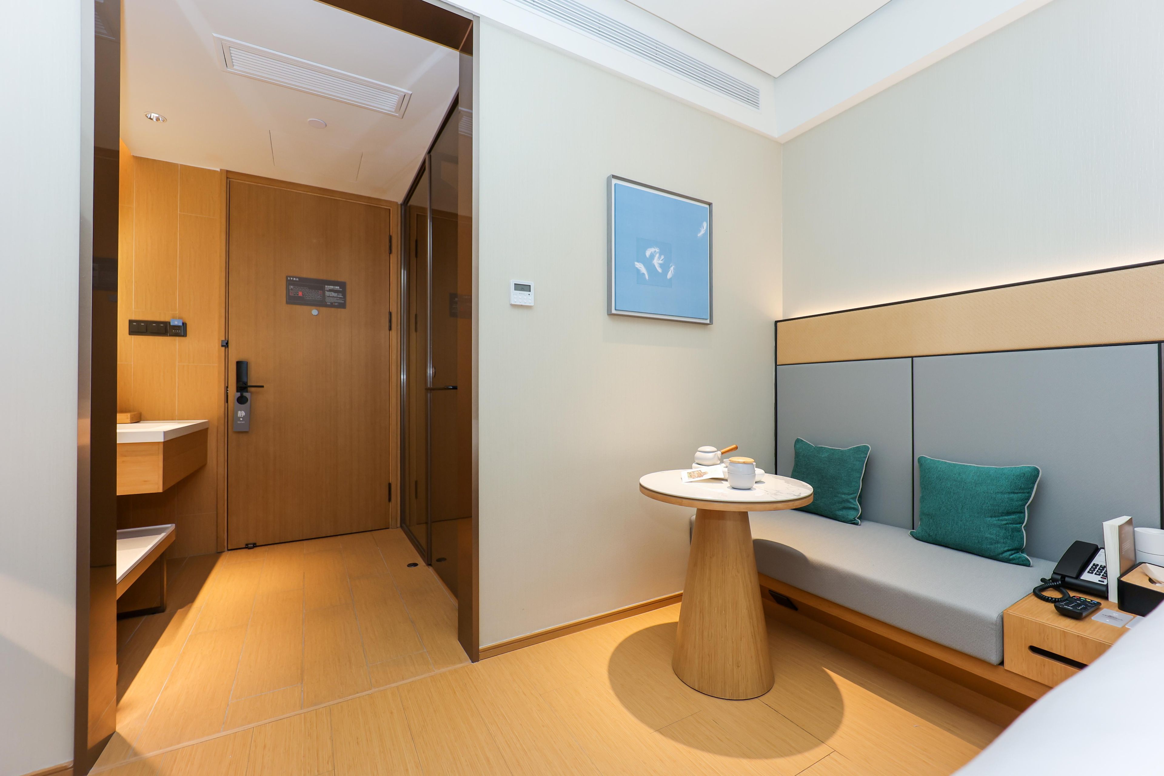 Ein modernes Hotelzimmer mit Holzboden, einem runden Tisch mit Teekanne, einer Couch mit Türkis-Kissen und einem an der Wand hängenden Kunstwerk.