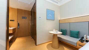A modern hotel room with a wooden floor, a round table with a teapot, a sofa with teal cushions, and a wall-mounted artwork.