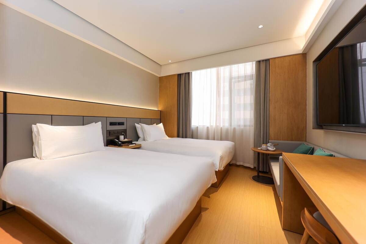 Quarto de hotel com duas camas, uma escrivaninha, uma cadeira, uma televisão e uma janela com cortinas.