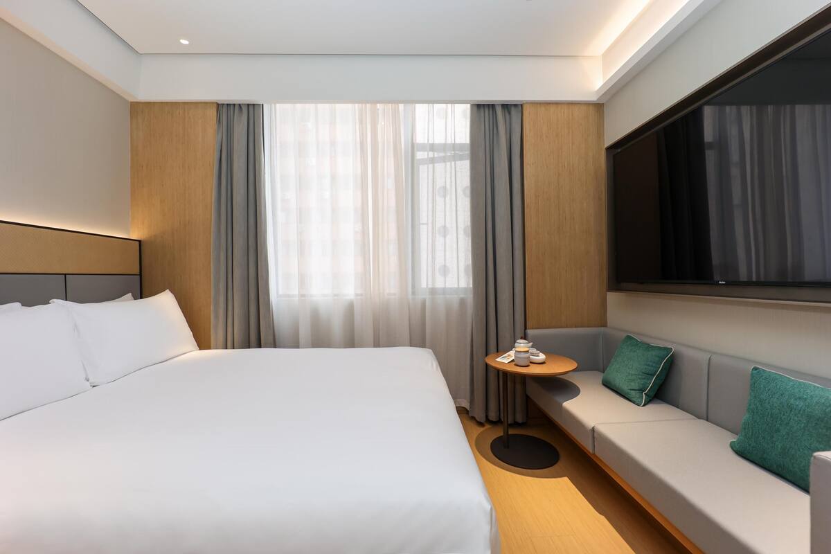 Quarto de hotel moderno com uma cama grande, um sofá, uma mesinha e uma televisão.