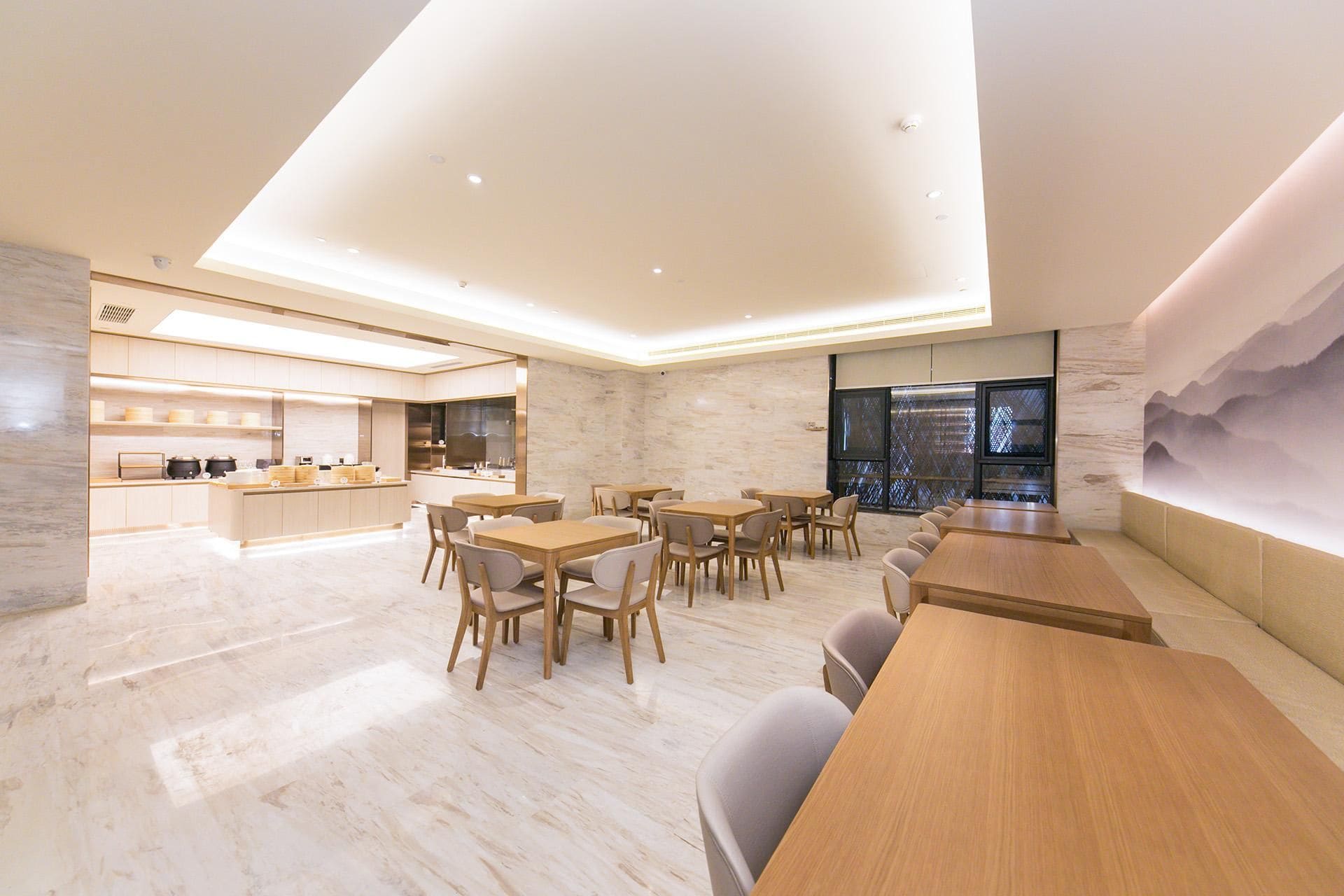 A modern dining area with wooden tables and chairs, a kitchen in the background, and a large window with a view.