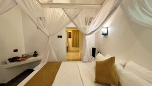 A bedroom with a bed, a nightstand, a wall-mounted speaker, and a doorway leading to another room. - Emerald Palm Courtyard Hotel (Zanzibar Town)