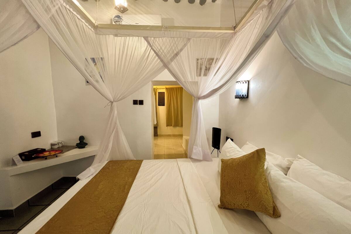 Un dormitorio con cama, mesita de noche, altavoz en la pared y un umbral que da a otra habitación.