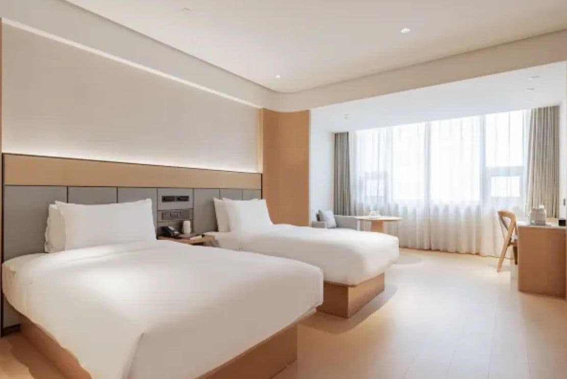 A hotel room with two beds, a small desk, a chair, and a large window with curtains.