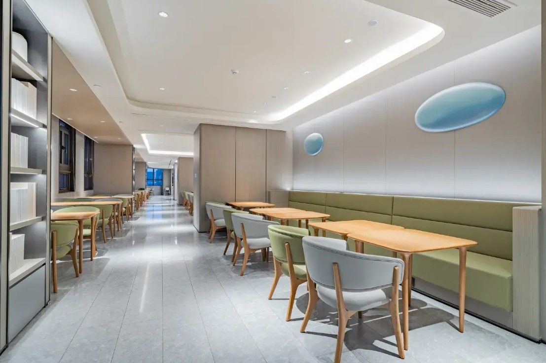 Ein modernes Interieur mit einer langen Reihe von Tischen und Stühlen, einer zentralen Sitzgruppe und einer Wand mit einem runden Designelement.