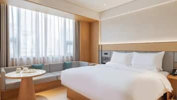 Una habitación de hotel moderna con una cama grande, una zona de estar con una mesa y un amplio ventanal con cortinas.