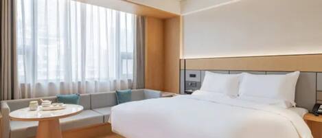 A modern hotel room with a large bed, a seating area with a table, and a large window with curtains.