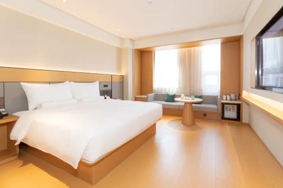 A modern hotel room with a large bed, a small seating area, and a window with curtains.