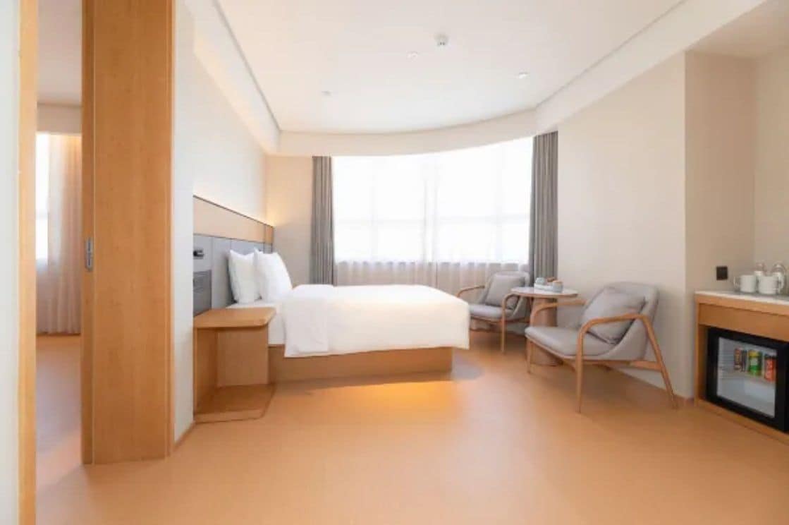 A modern hotel room with a bed, bedside table, chair, and a small table with a refrigerator.