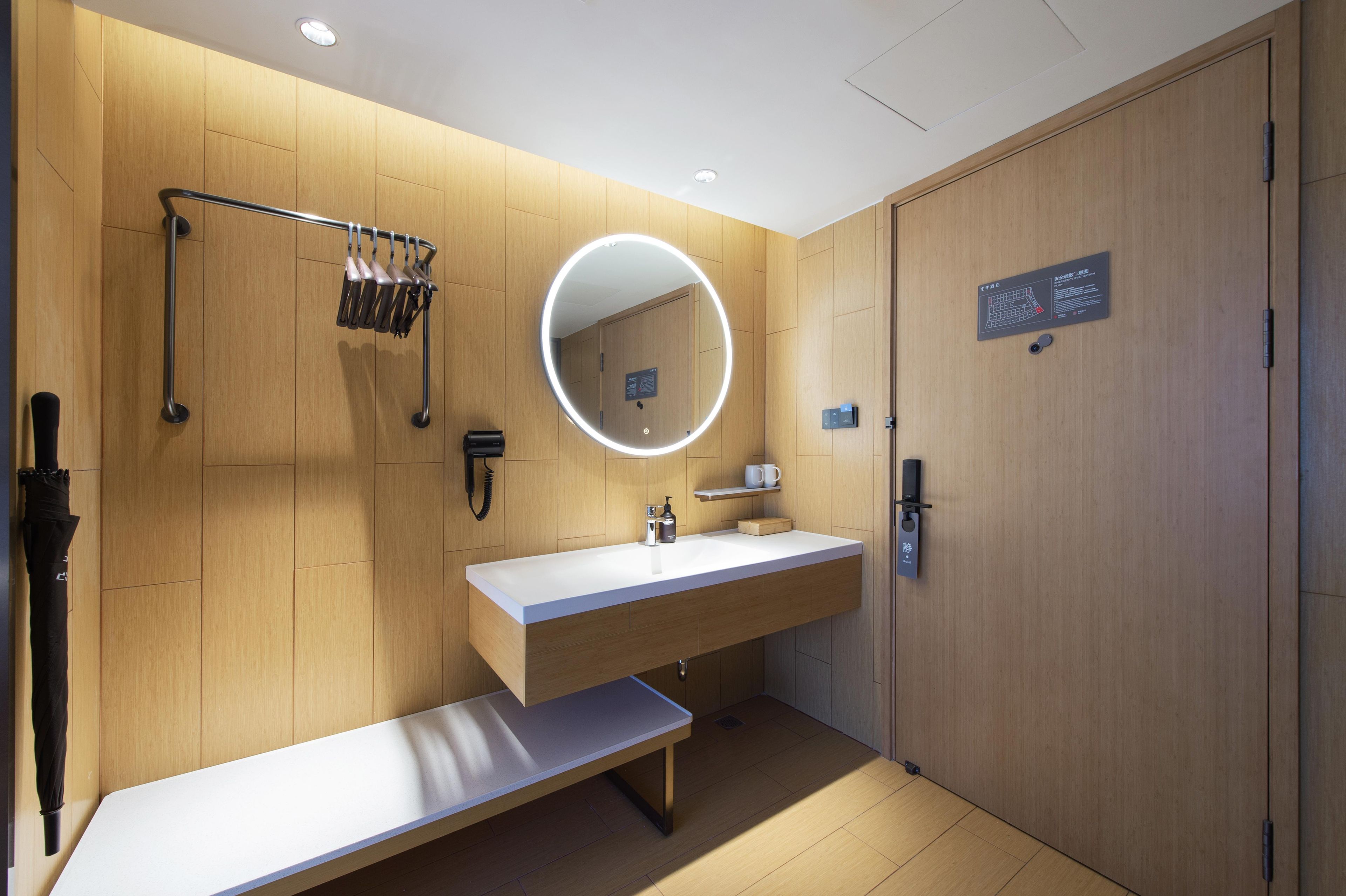 A modern bathroom with a wooden vanity, a round mirror with lighting, a bench, and a towel rack.