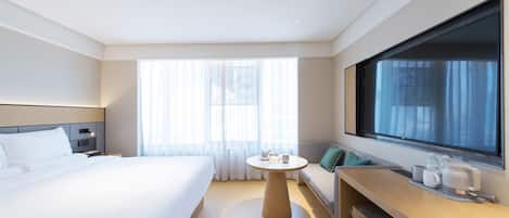 Habitación de hotel con cama, una mesa redonda con cafetera, un sofá y un ventanal grande.