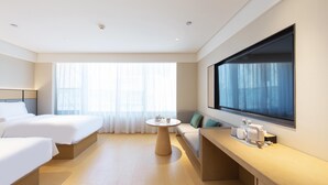 Ein Hotelzimmer mit zwei Betten, einem runden Tisch, einer Couch, einem großen Fenster und einem flachbildfernseher, der an der Wand befestigt ist.