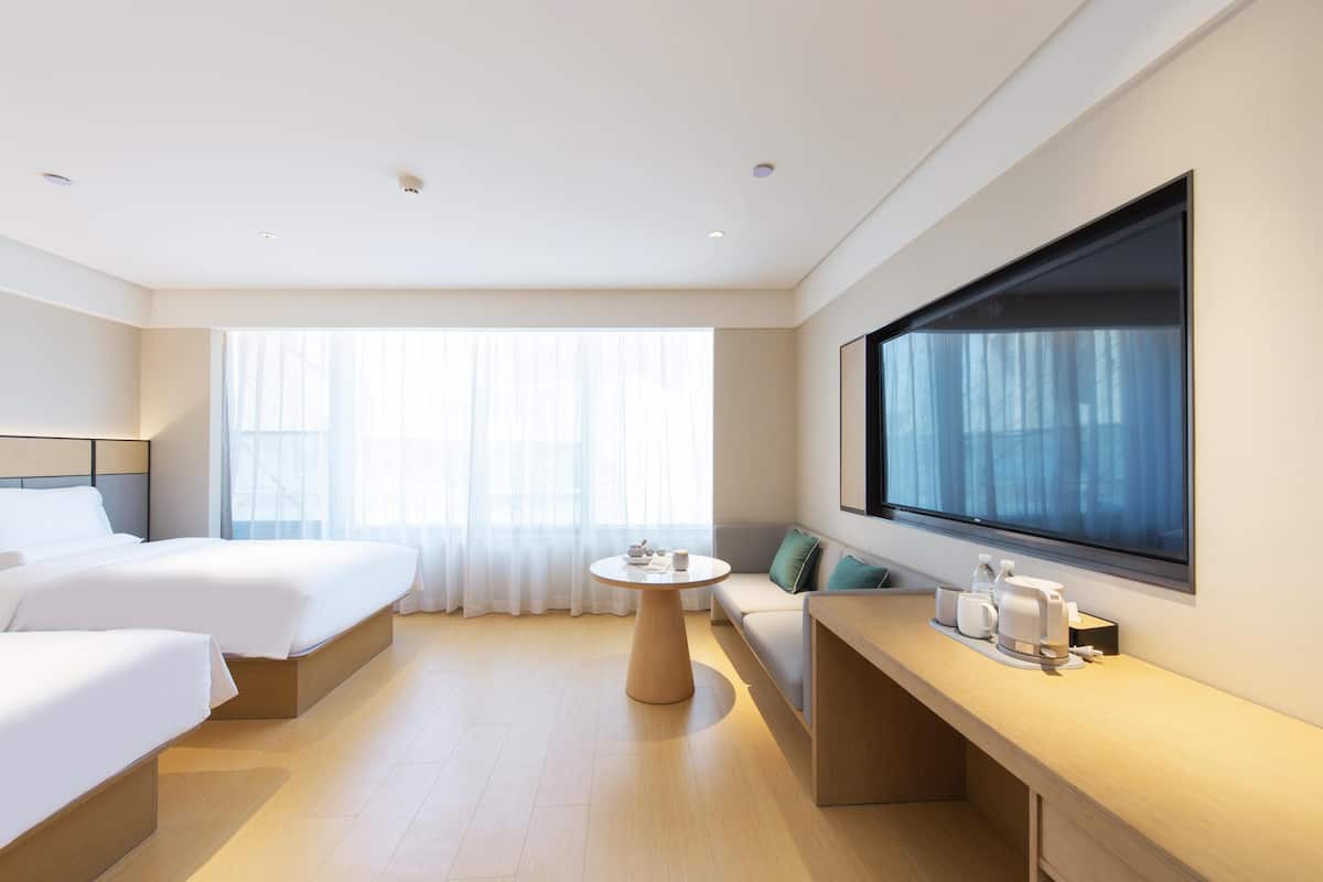 Habitación de hotel con dos camas, una mesa redonda, un sofá, un ventanal grande y un televisor de pantalla plana montado en la pared.