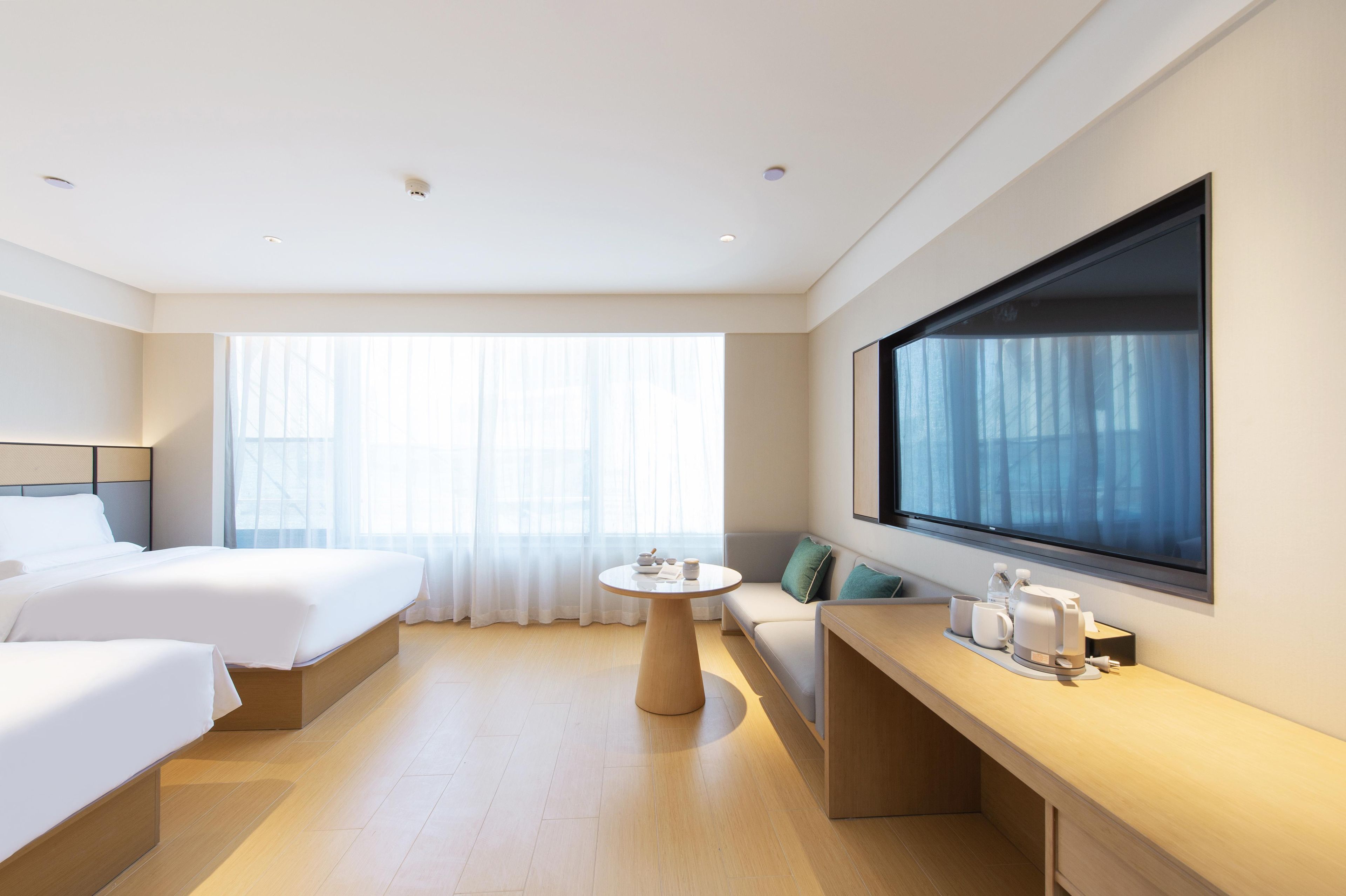 A hotel room with two beds, a round table, a sofa, a large window, and a flat-screen TV mounted on the wall.