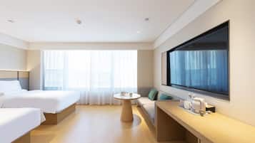 A hotel room with two beds, a round table, a sofa, a large window, and a flat-screen TV mounted on the wall.
