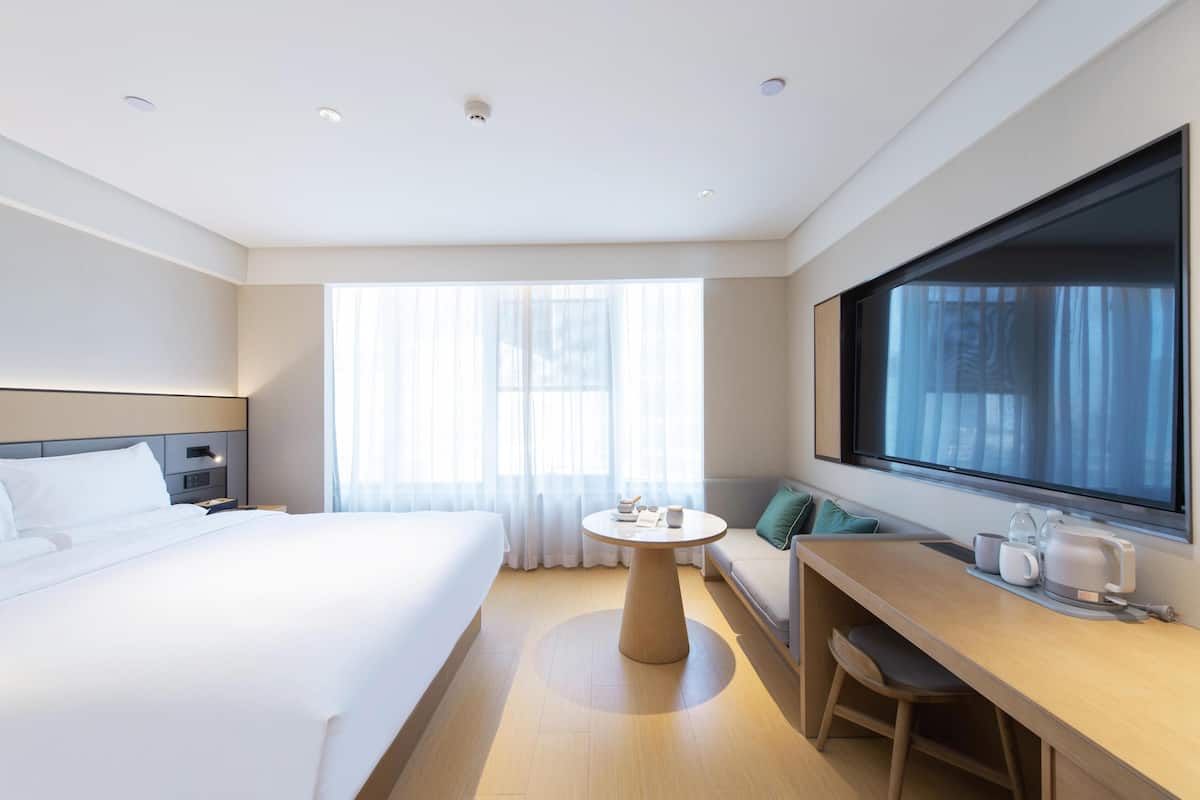 Habitación de hotel con cama, una mesa redonda con cafetera, un sofá y un ventanal grande.
