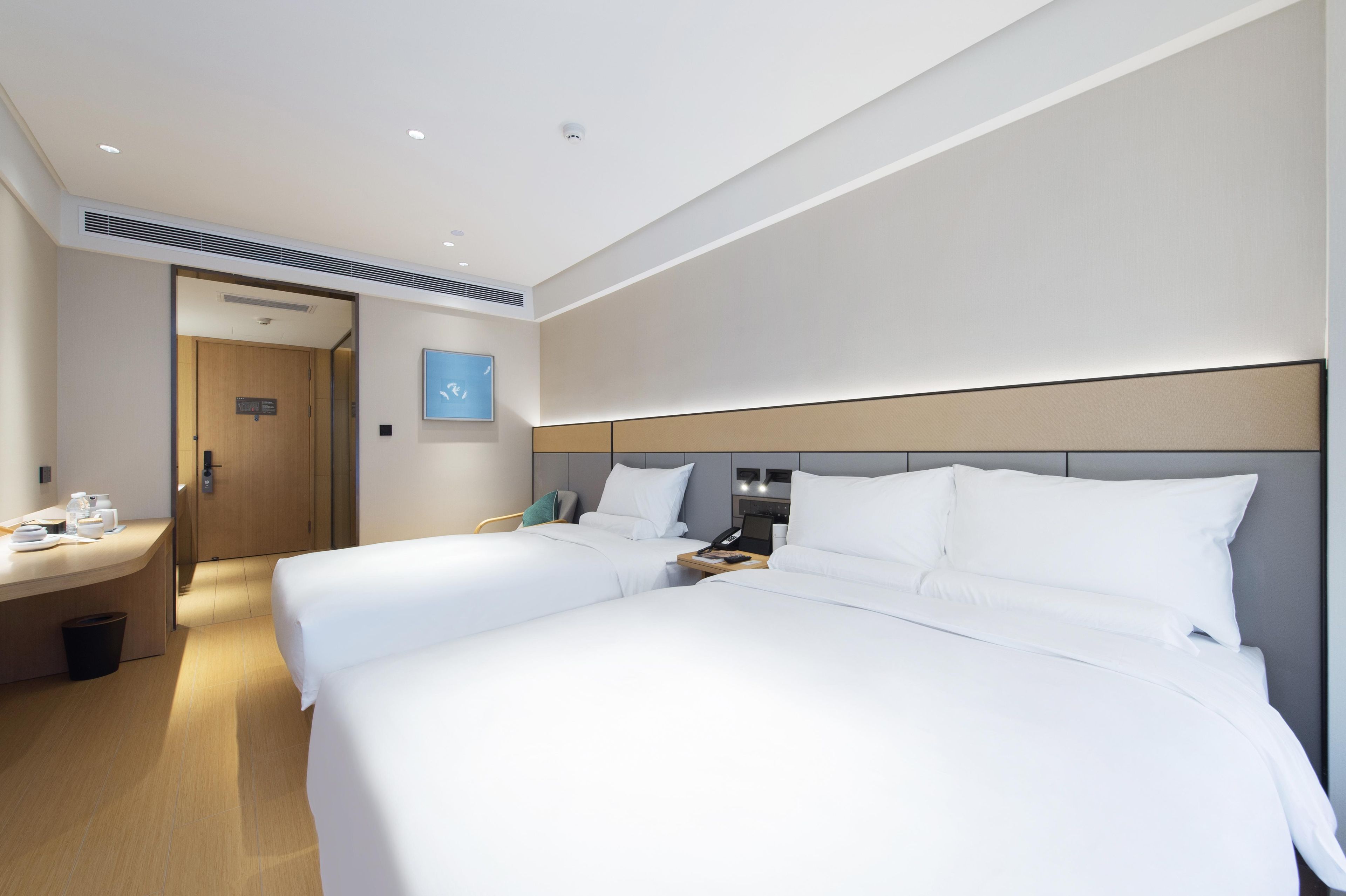 A hotel room with two beds, a desk, a chair, a trash bin, and a door leading to another room.