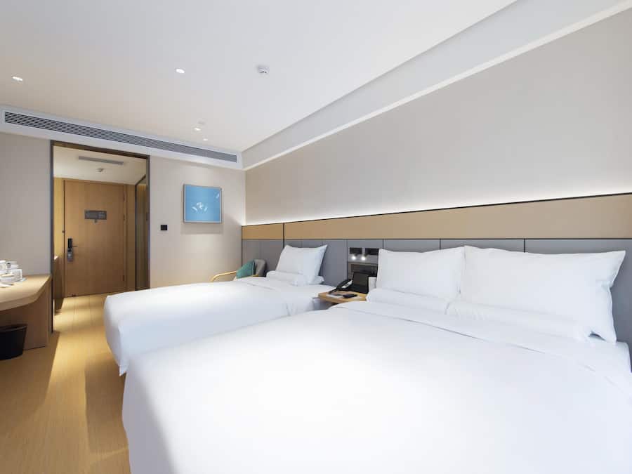 A hotel room with two beds, a desk, a chair, a trash bin, and a door leading to another room.
