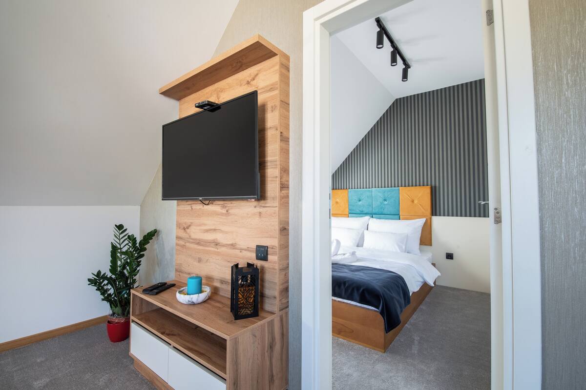 Un dormitorio con cama, televisión montada en una pared de madera, mesita de noche y planta en maceta.