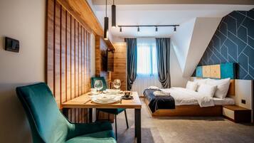 A modern hotel room with a wooden accent wall, a dining area with a table and chairs, a bed with a headboard, and a geometric-patterned wall.