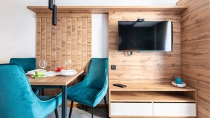 A modern room with a wooden wall, a flat-screen TV mounted above a wooden shelf, a dining table with chairs, and a small seating area. - Hotel Djina (Raška)