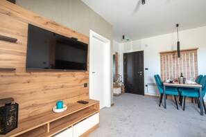 A modern living room with a flat-screen TV mounted on a wooden wall, a dining area with teal chairs, and a black door in the background.