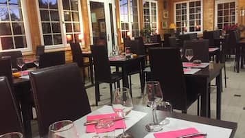 Um restaurante com mesas postas para refeições, cadeiras e vista para o exterior através de grandes janelas.