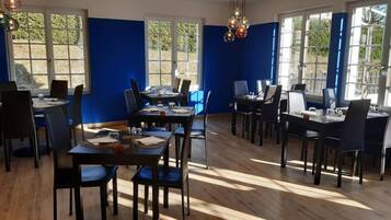 A dining area with multiple tables and chairs, large windows with a view of greenery, and a wooden floor.